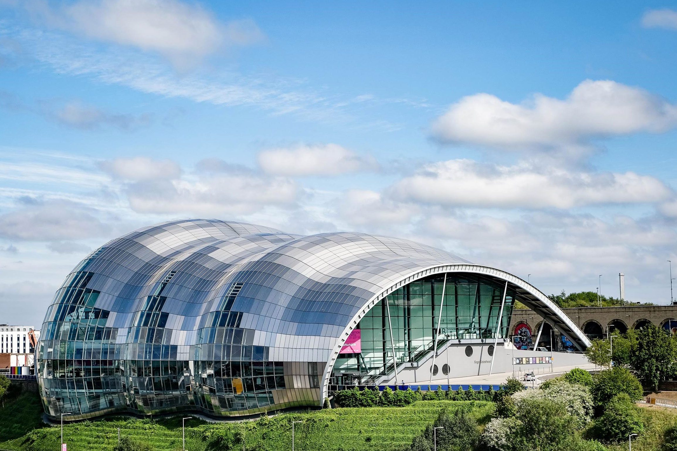 A view of The Glasshouse on a grassy slope under a blue sky with scattered clouds - picture taken from The Tyne Bridge.
