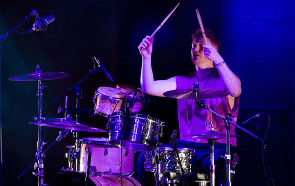 Artist Benjamin Fitzgerald holding drum sticks in the air at a drum kit in a darkened Sage Two