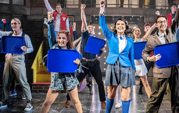 musical cast in checked skirts and blue jackets punching the air