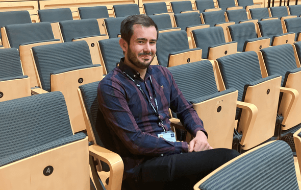 a person in a checked shirt with brown hair sitting on wood and blue chairs