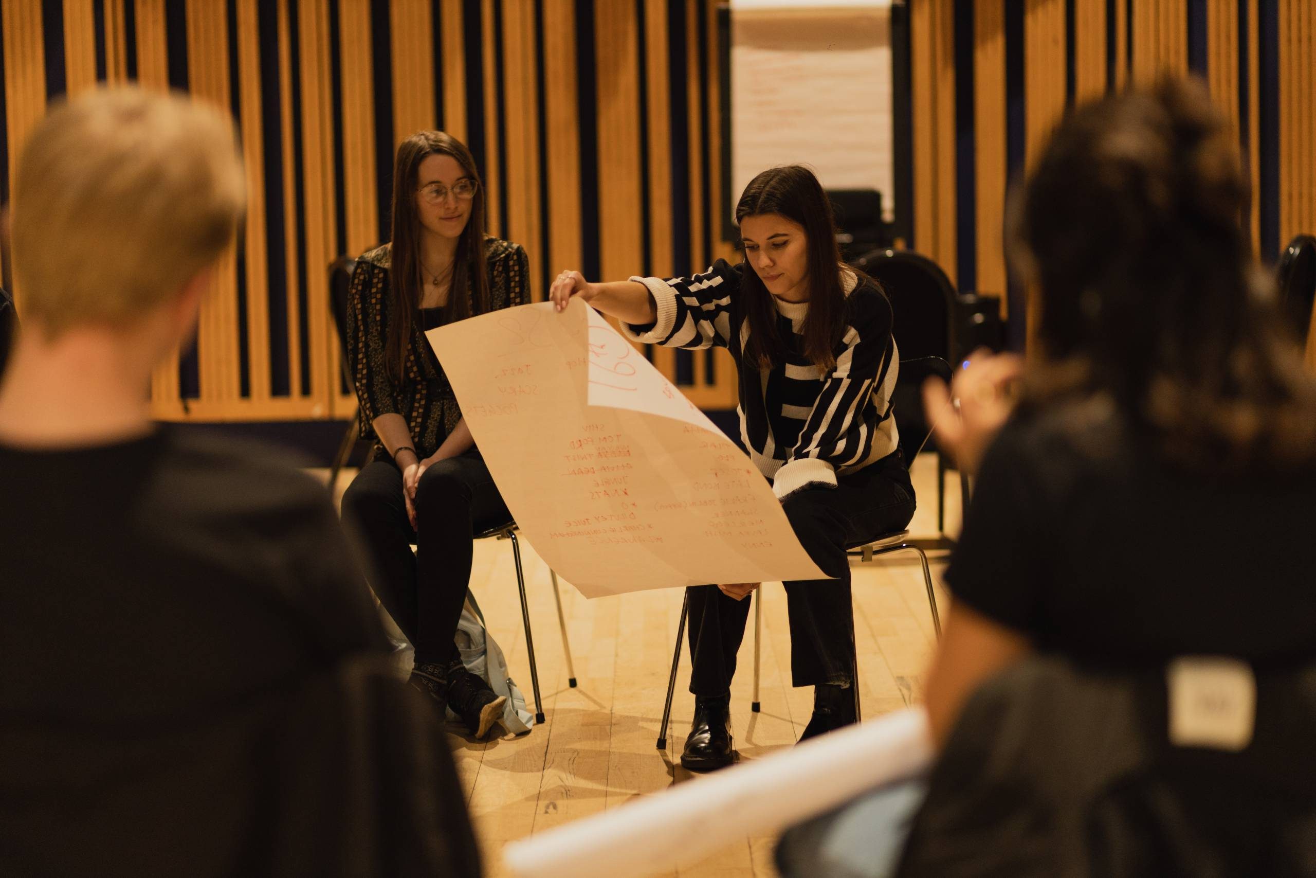 two people on chairs, one is holding a big piece of white paper