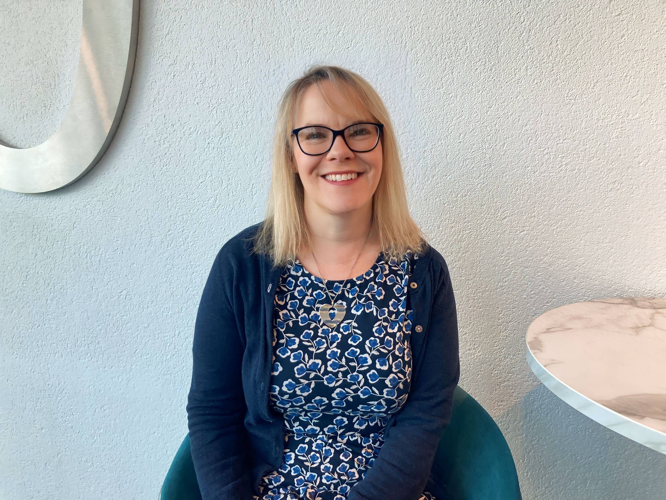 person with blonde hair, glasses and a blue dress sitting