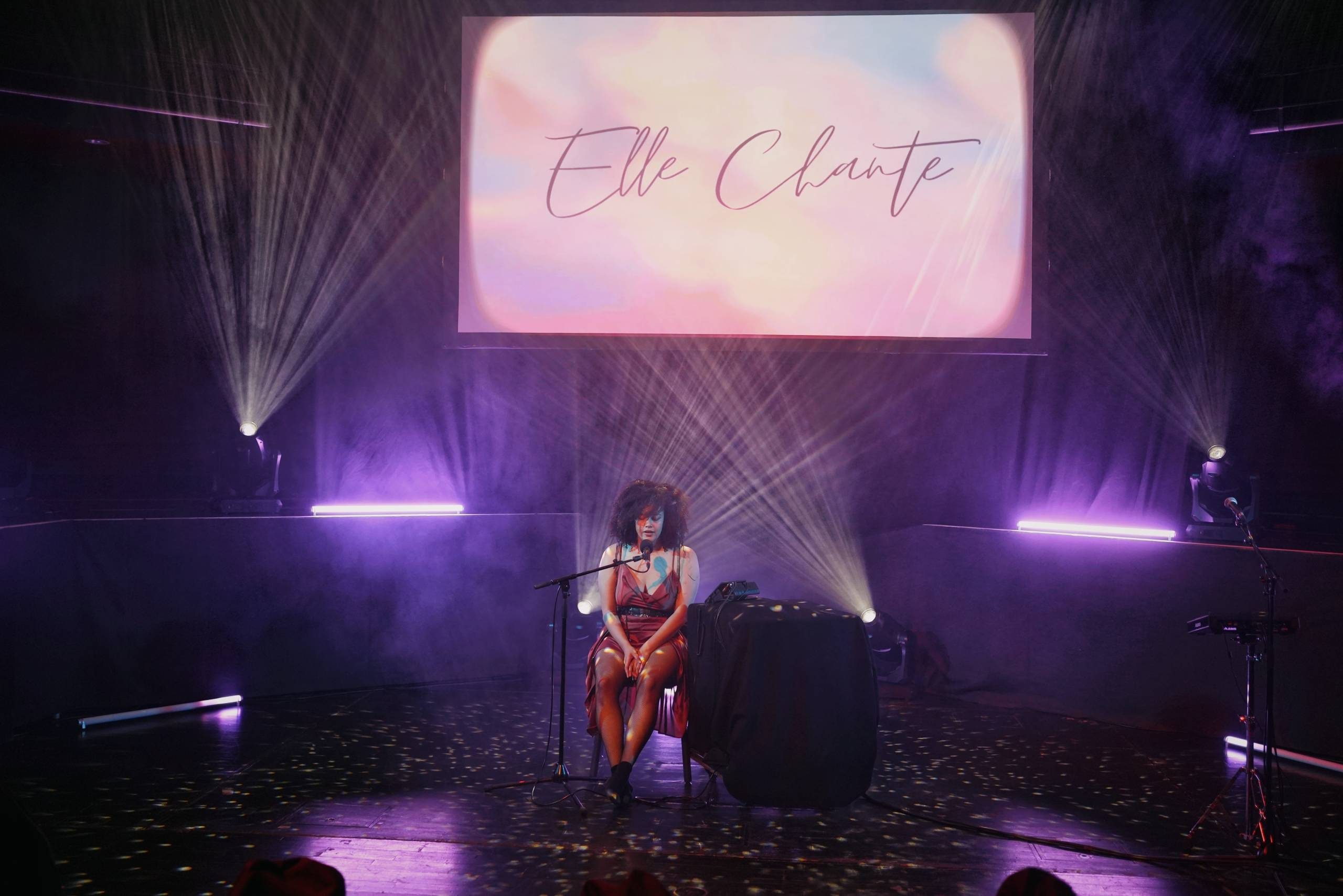 A woman with natural dark brown curly hair wearing a reddish brown dress and a sparkly black belt. She is sitting on a chair and singing into a microphone on a stage, which has purple and and white lighting plus a screen with Elle Chante's logo and with a background that looks like a sunrise in the background.