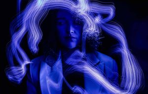 A Caucasian woman with shoulder length curly hair bathed in purple light. She is wearing a pale tailored blazer with a black top underneath and has a septum piercing. There are abstract purple lines surrounding her.