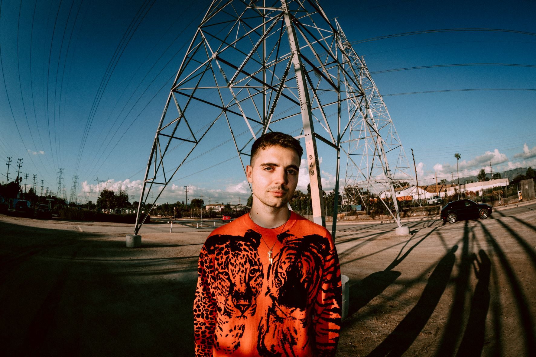 A white male with short brown hair wears a brightly coloured jumper with large black prints of a leaopard and a tiger on it. He is stood at the foot of a large electricity pylon by a road in an urban environment. The camera uses a fish-eye lens to fit a wider perspective. There is bright blue sky across the top half of the photo.