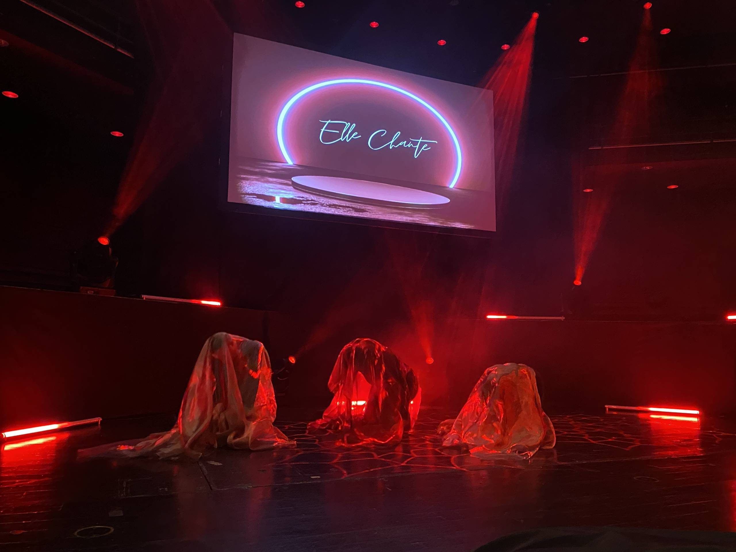 Trio of dancers under red lights and white sheets. There is a screen with a logo above them.