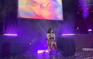 A person in a colourful dress and black hair sitting on a stool under purple lights and a colour-filled screen