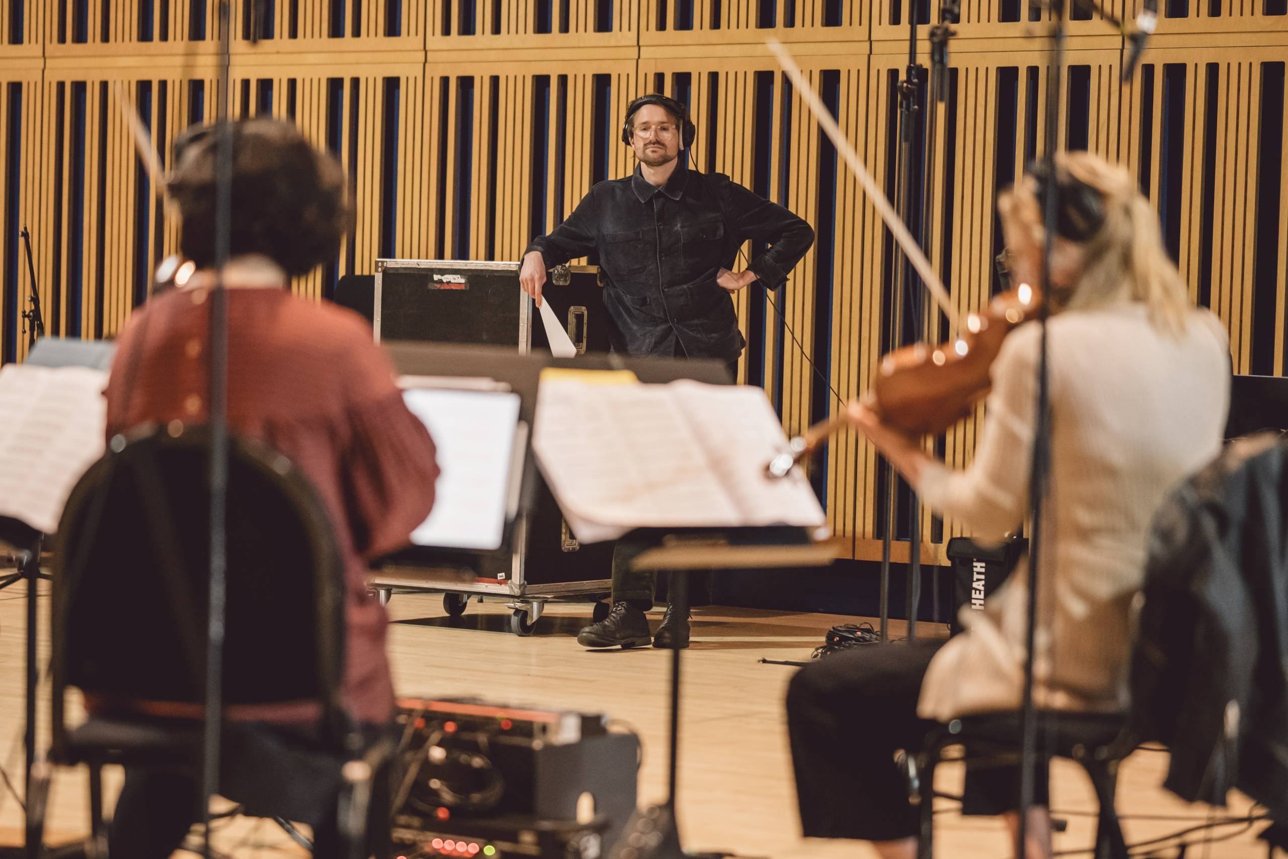 Hayden Thorpe, a white man in his thirties with dark hair, wearing glasses and a dark shirt, watches as two seated violin players play during a recording session.