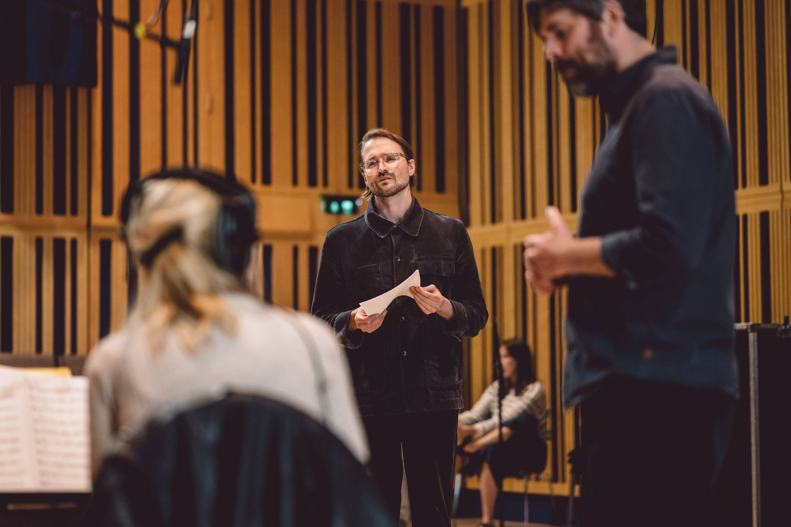Hayden Thorpe, a white man in his thirties with dark hair, wearing glasses and a dark shirt, holds a sheet of music during a conversation between music arranger Jack McNeil (who is a white male with a dark beard and hair) and a seated musician with tied back blonde hair, who is wearing headphones.