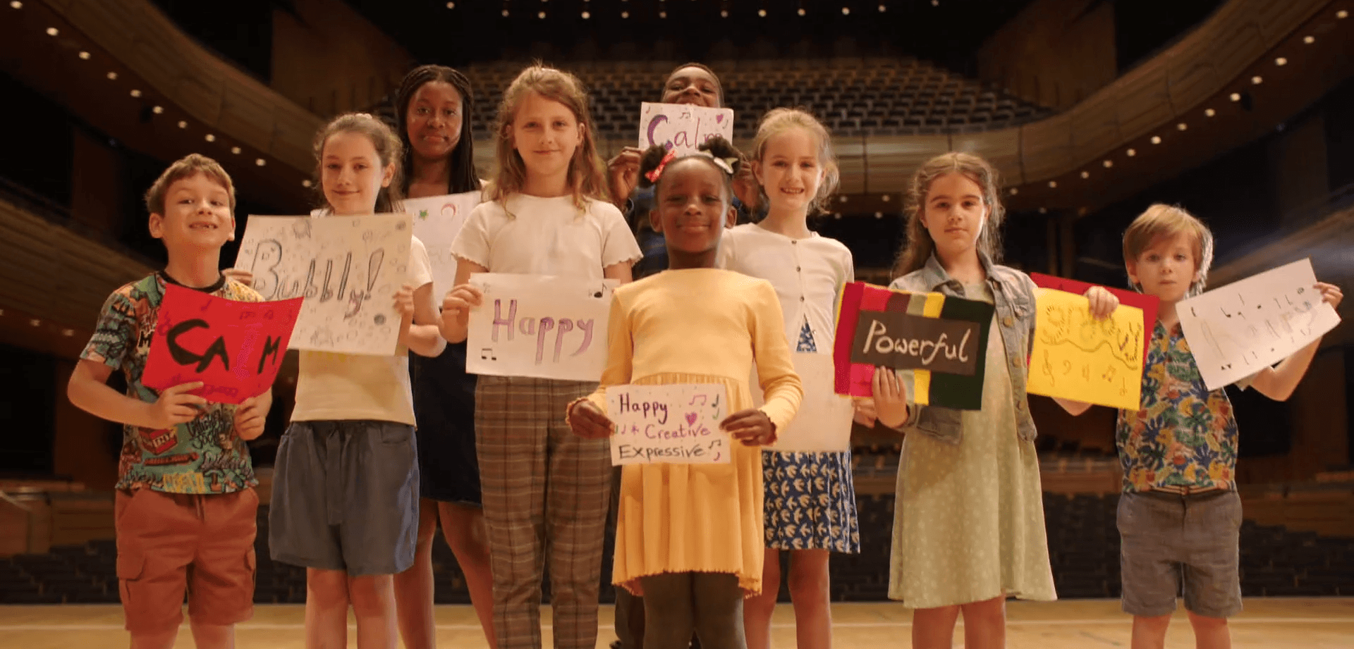 A group of smiling young children holding cards with words like happy, powerful, bubbly and joyful to describe how music makes them feel.