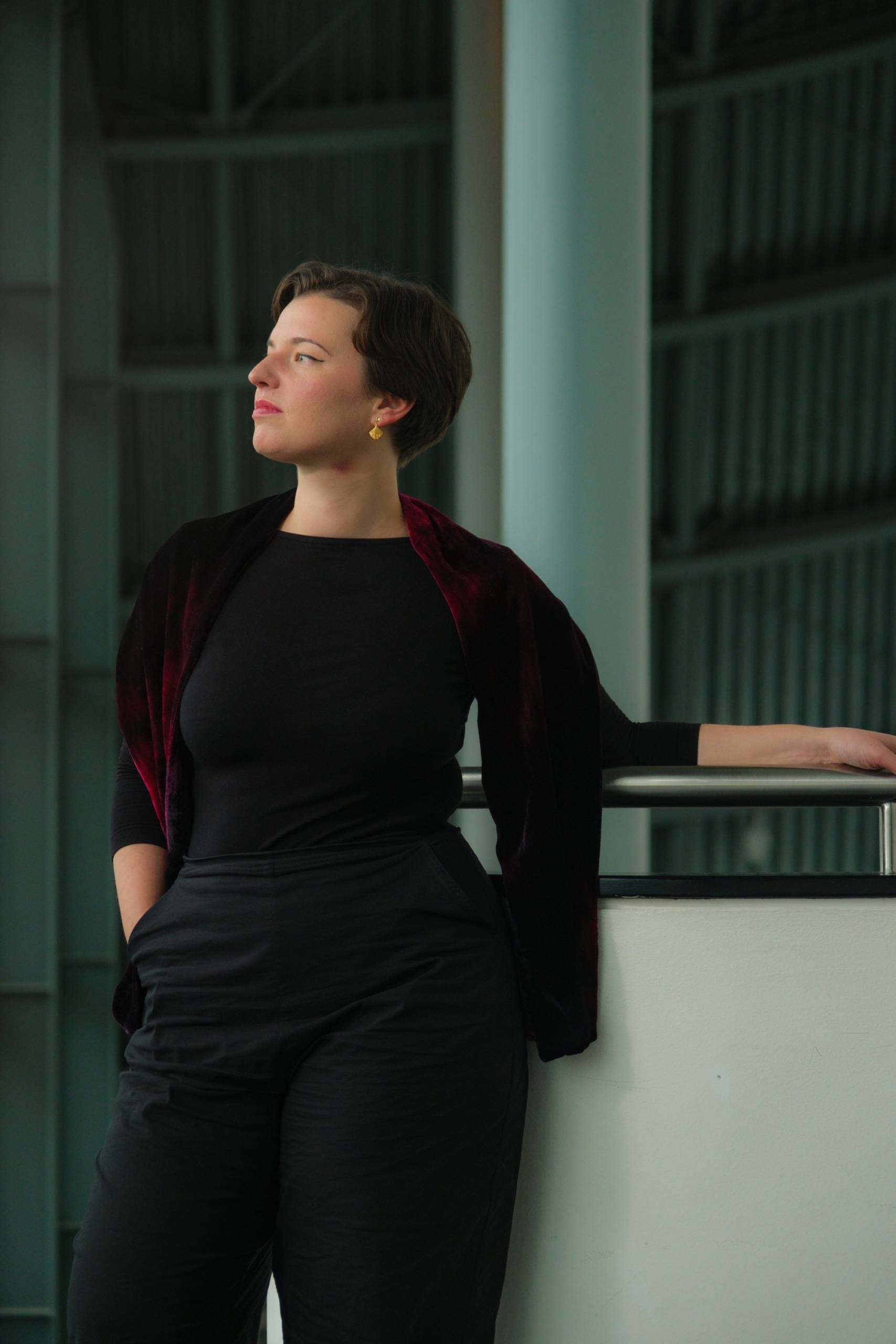 Maria Włoszczowska at The Glasshouse. Maria - a white female in her 30s is leaning against the top of a staircase in the building concourse. She is wearing black trousers and top, with a dark red throw around her shoulders. She is looking away from the camera, out of a window to the left of the frame.