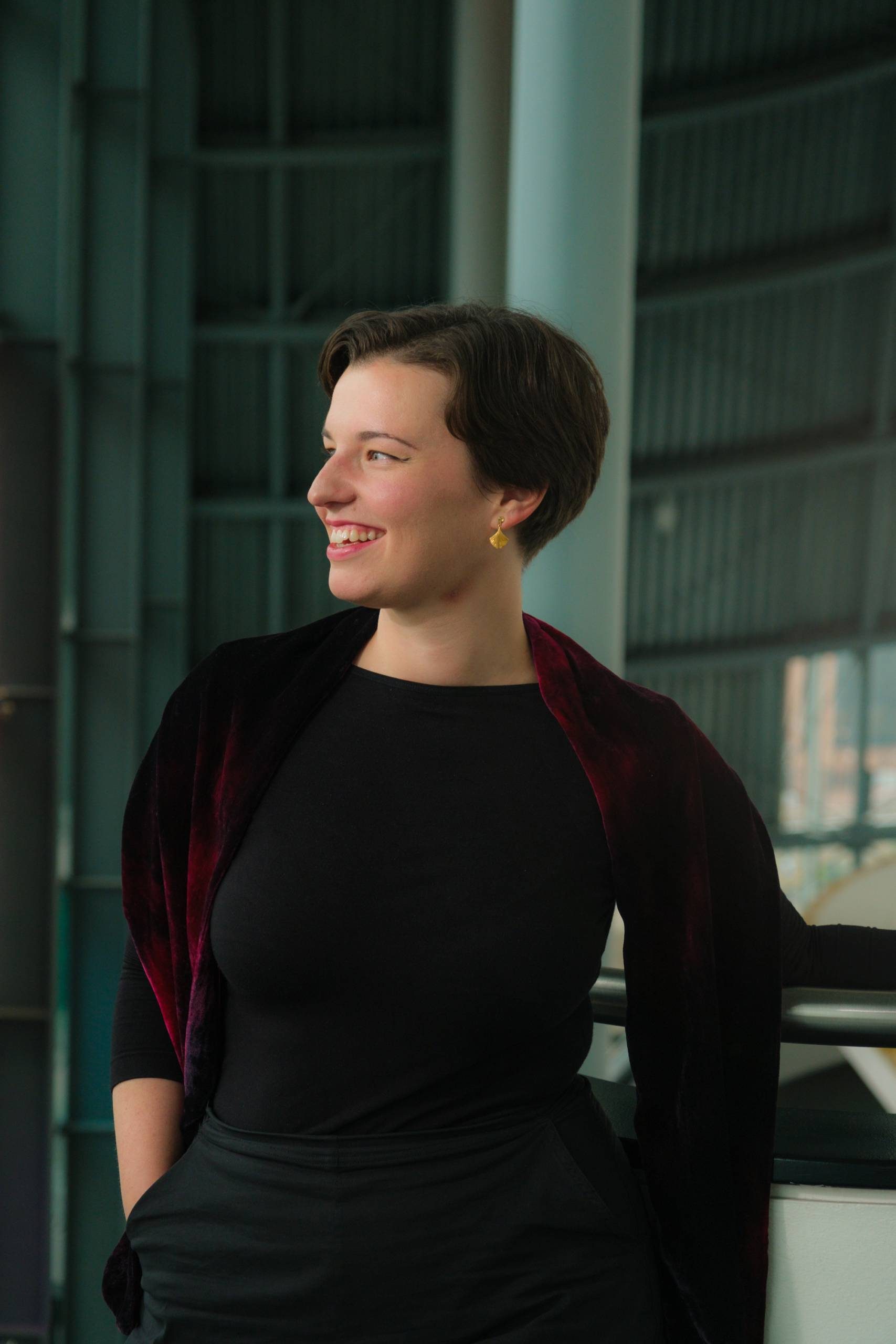 Maria Włoszczowska at The Glasshouse. Maria - a white female in her 30s is leaning against the top of a staircase in the building concourse. She is wearing black trousers and top, with a dark red throw around her shoulders. She is smiling, looking away from the camera, out of a window to the left of the frame.