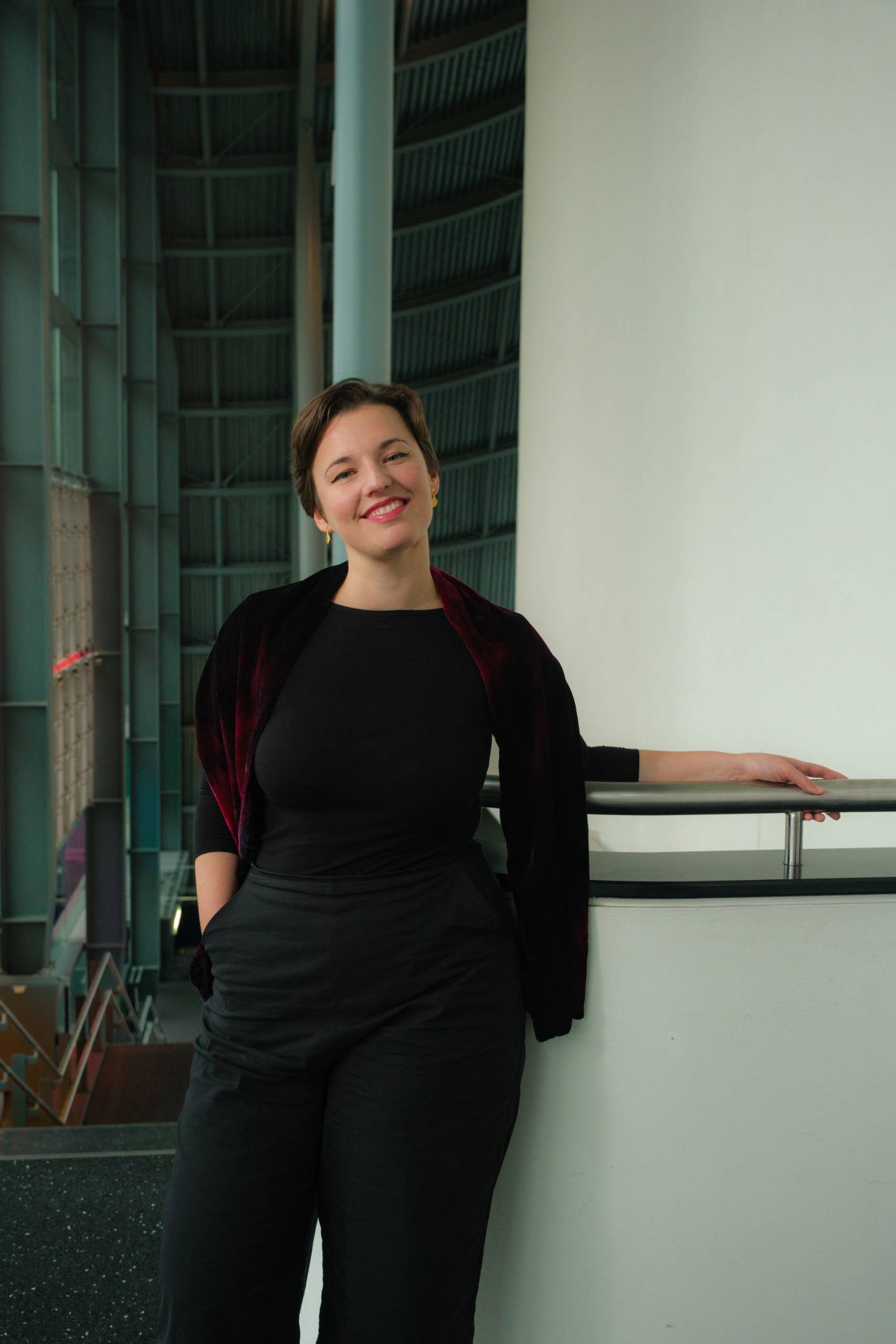 Maria Włoszczowska at The Glasshouse. Maria - a white female in her 30s is leaning against the top of a staircase in the building concourse. She is wearing black trousers and top, with a dark red throw around her shoulders. The photo frames Maria from the waist up, and she is smiling happily, looking straight at the camera.