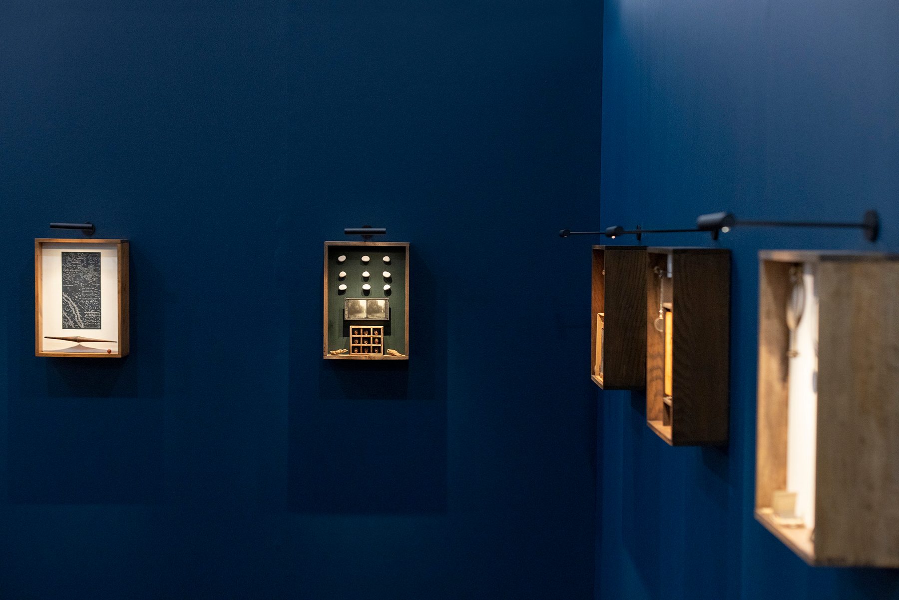 Wooden display boxes with artwork and objects are mounted on a deep blue wall, each box lit from above by a small spotlight.