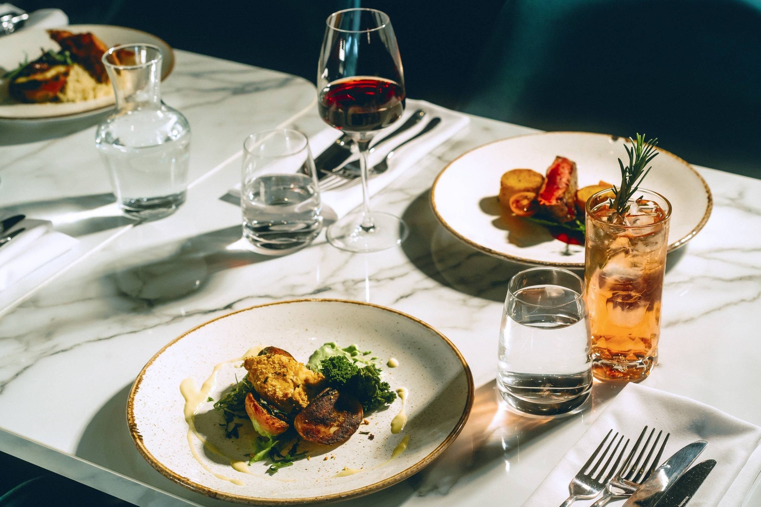 Plated gourmet meals and drinks set on a marble-top table, with glasses of water, red wine, and a cocktail garnished with rosemary.
