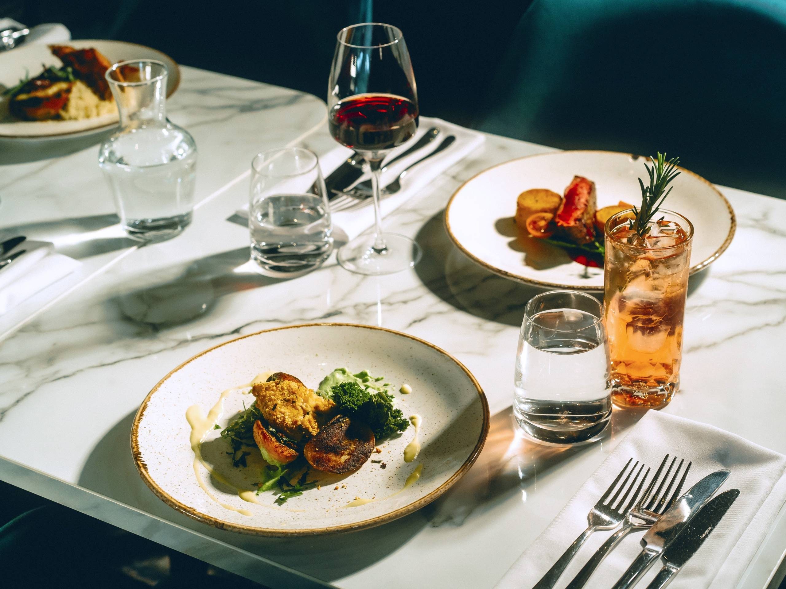 Plated gourmet meals and drinks set on a marble-top table, with glasses of water, red wine, and a cocktail garnished with rosemary.