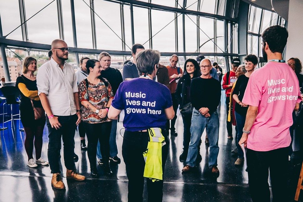A group of people stand in the Concourse of The Glasshouse, listening attentively to two tour guides wearing shirts that say Music lives and grows here.