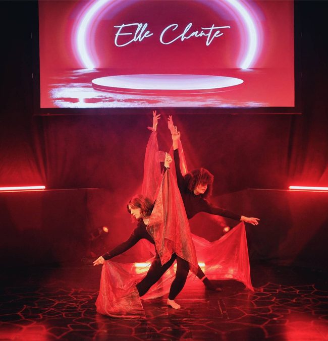 Two dancers in black outfits perform with flowing red fabric on a dim Sage Two stage, illuminated by red lights.