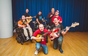 A group of smiling happy musicians in a rehearsal room, some are holding guitars and one is playing on a drumkit.