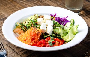 A white bowl filled with colorful vegetables sits on a wooden table next to a drink and a fork.
