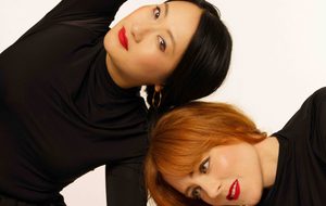 Hannah Peel and Beibei Wang: Two women wearing black clothes and red lipstick. Their hair is braided together.