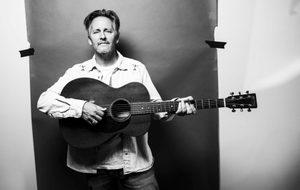 Josh Rouse holding an acoustic guitar in a black and white portrait.