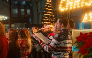 Singers in festive jumpers performing together beside a Christmas tree at The Glasshouse.