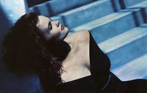 Belinda Carlisle reclining in a black outfit, looking upwards against a blue background.