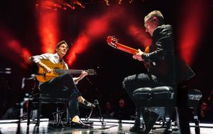Two men perform on a stage, holding guitars with red lighting
