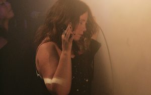 A woman in a black dress sings into a microphone on a dim, smoky stage, raising one hand by her face