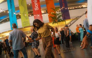 A woman with dark hair wearing brown trousers and a yellow Fred Perry Polo shirt dances with other people in the background on The Concourse at The Glasshouse.