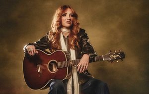 Kezia Gill: A woman with long auburn hair wearing dark blue jeans, a brown leather jacket, a white scarf and boots sits with an acoustic guitar in her lap.