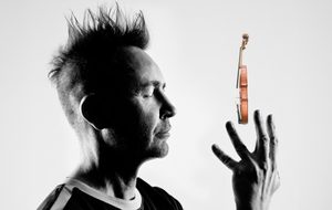 Black-and-white portrait of a spiky-haired man looking towards at a small floating violin above his raised hand with his eyes closed