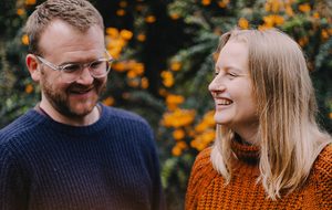 A smiling duo in navy and orange sweaters laugh outdoors, surrounded by blurred orange flowers and green foliage.