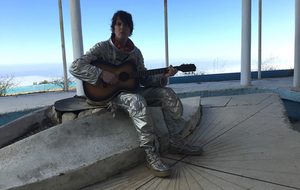 A person in a metallic space suit sitting on a concrete platform and playing an acoustic guitar.