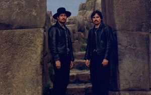 Two brothers stand side by side with a staircase behind them.